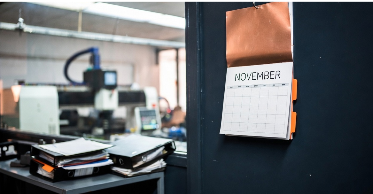 November calendar above a desk of compliance binders in a quiet precision machine shop office, with CNC equipment visible through the window.